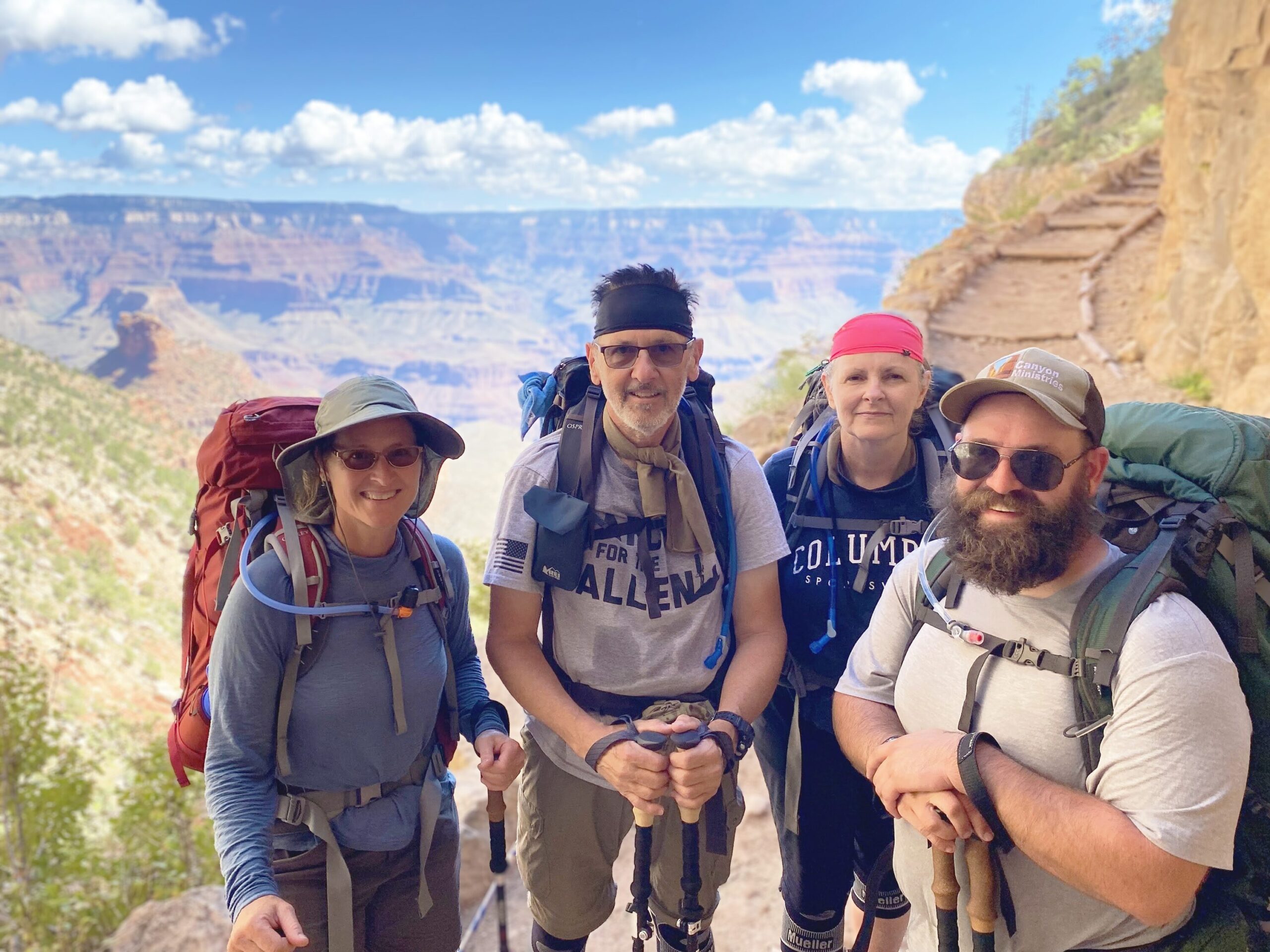 Bryce and Dana on Grand Canyon Backpacking Trip REI Adventures Four hikers on a backpacking trip stand on a trail in the Grand Canyon, smiling for a photo. They carry backpacks and wear hats and sunglasses. The canyon and a bright sky with scattered clouds are visible in the background.