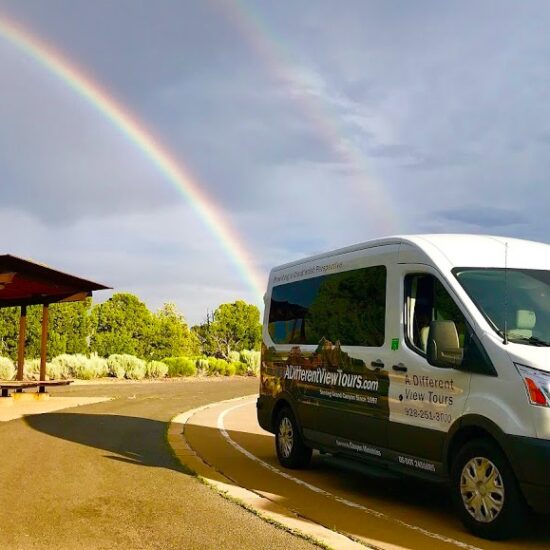 A white tour van from Canyon Ministries is parked by a rest area with a wooden shelter. A vibrant double rainbow arcs across the partly cloudy sky, surrounded by lush green trees and shrubs.